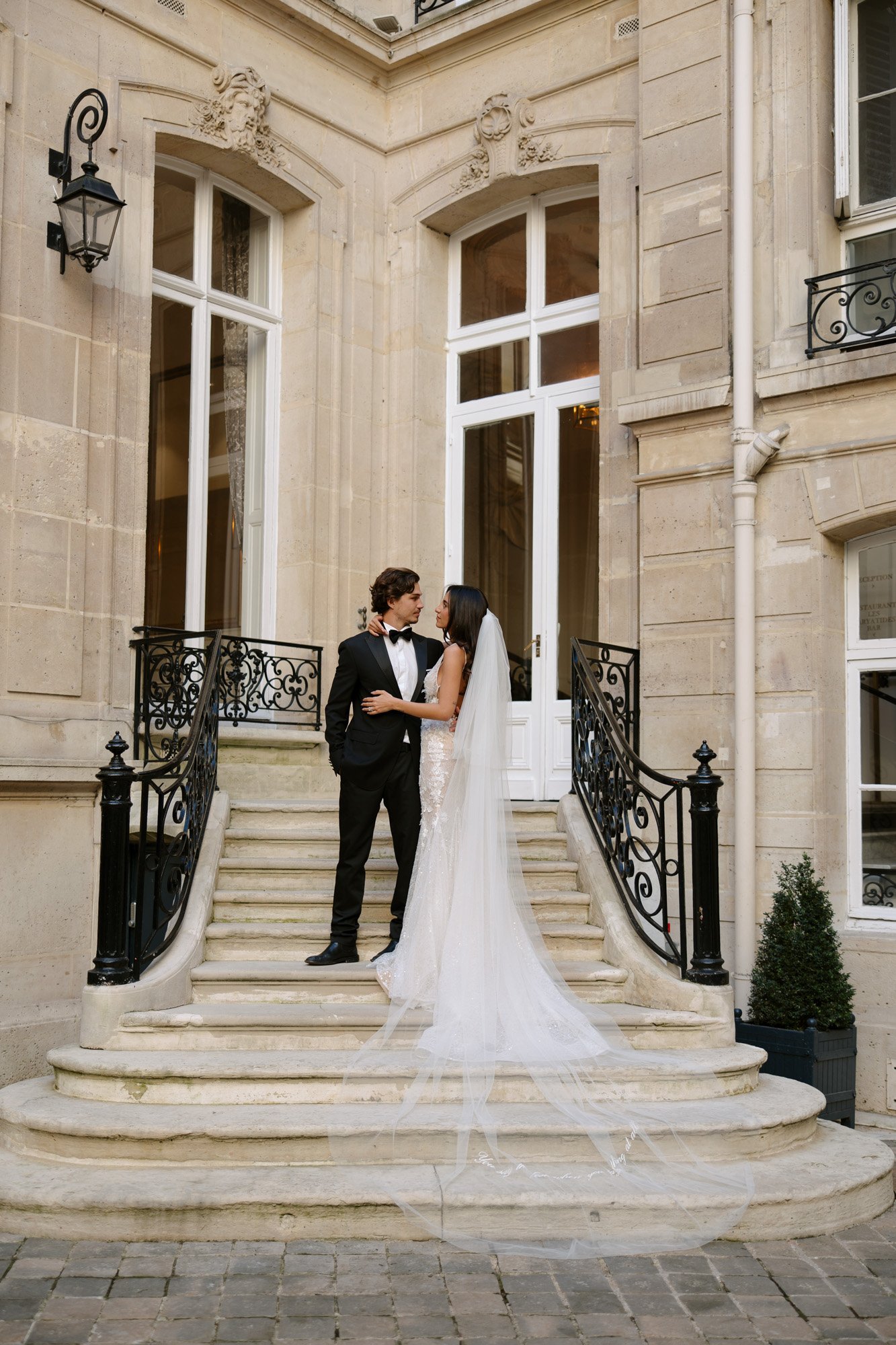 A bride and groom stand together on the steps of a grand stone building, facing each other and holding hands, with the bride wearing a long white dress and veil. Paris wedding photographer.