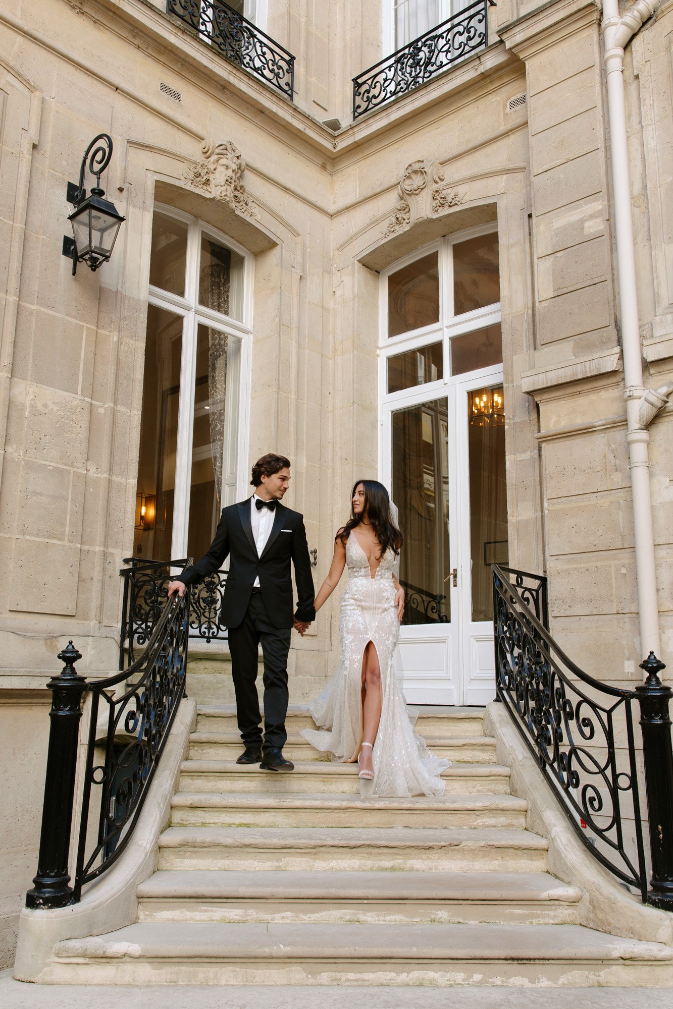 A man in a black tuxedo and a woman in a white gown walk down stone steps outside an elegant building with large windows and wrought iron railings. Hotel Alfred Sommier Elopement.