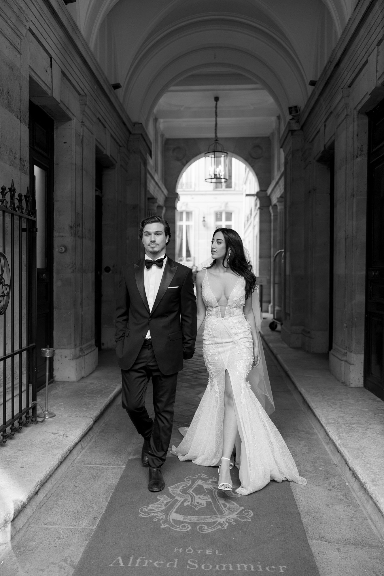 A man in a tuxedo and a woman in a wedding dress walk side by side down a covered stone corridor at Hôtel Alfred Sommier.