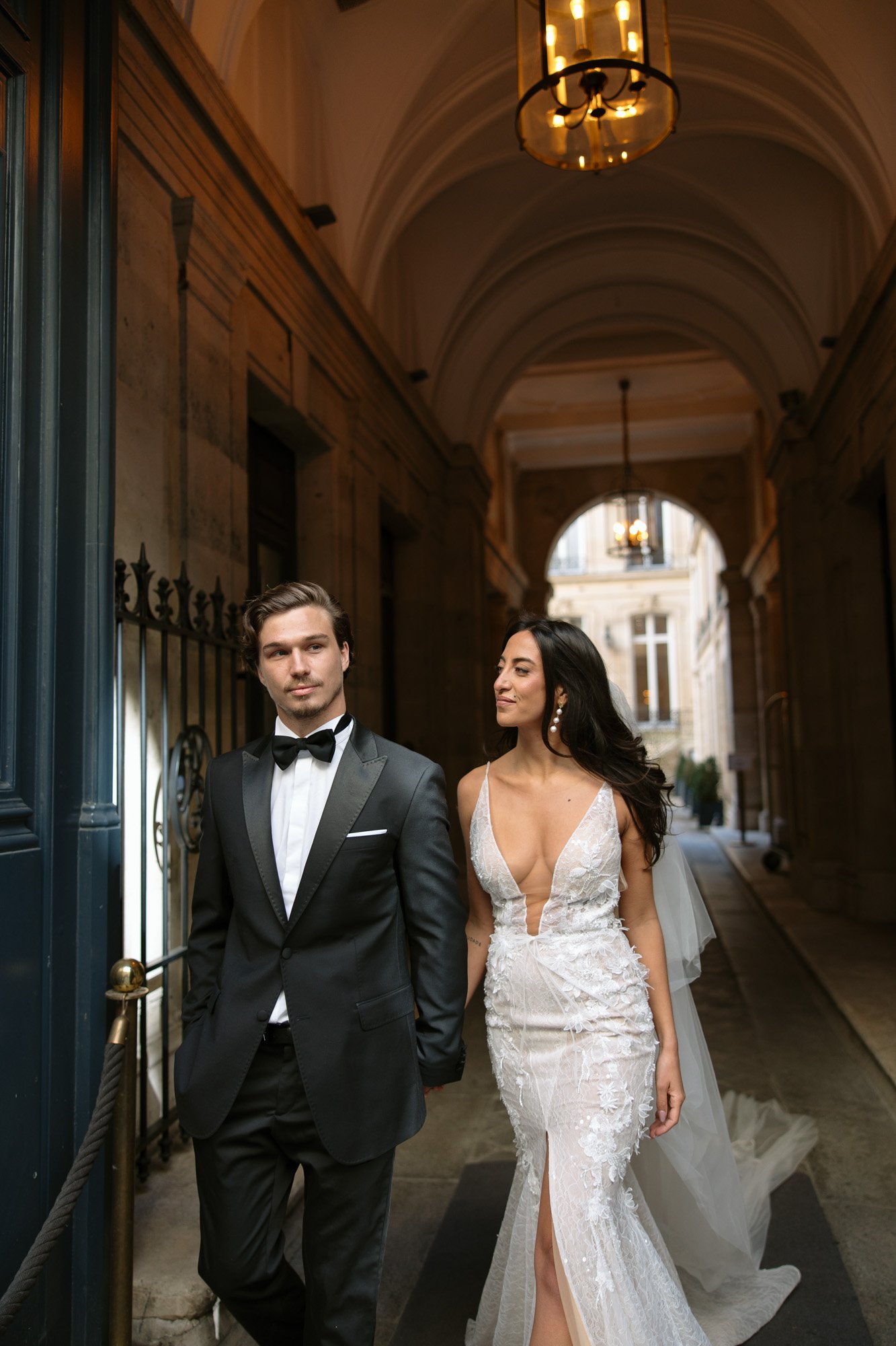 A man in a black tuxedo and a woman in a white, sleeveless wedding dress walk together in a grand, arched corridor with elegant lighting.  Hotel Alfred Sommier Elopement.