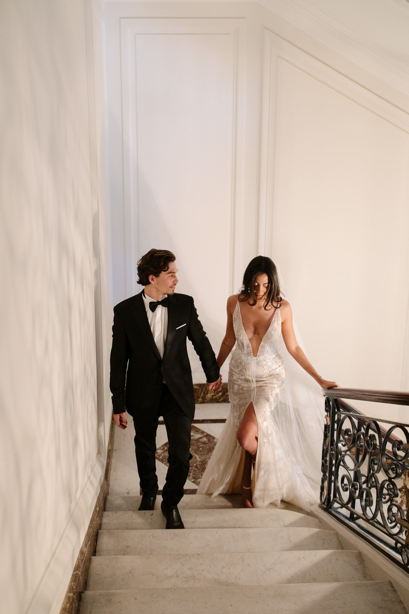 A man in a black tuxedo and a woman in a white wedding dress walk down a staircase, holding hands. Hotel Alfred Sommier Elopement.