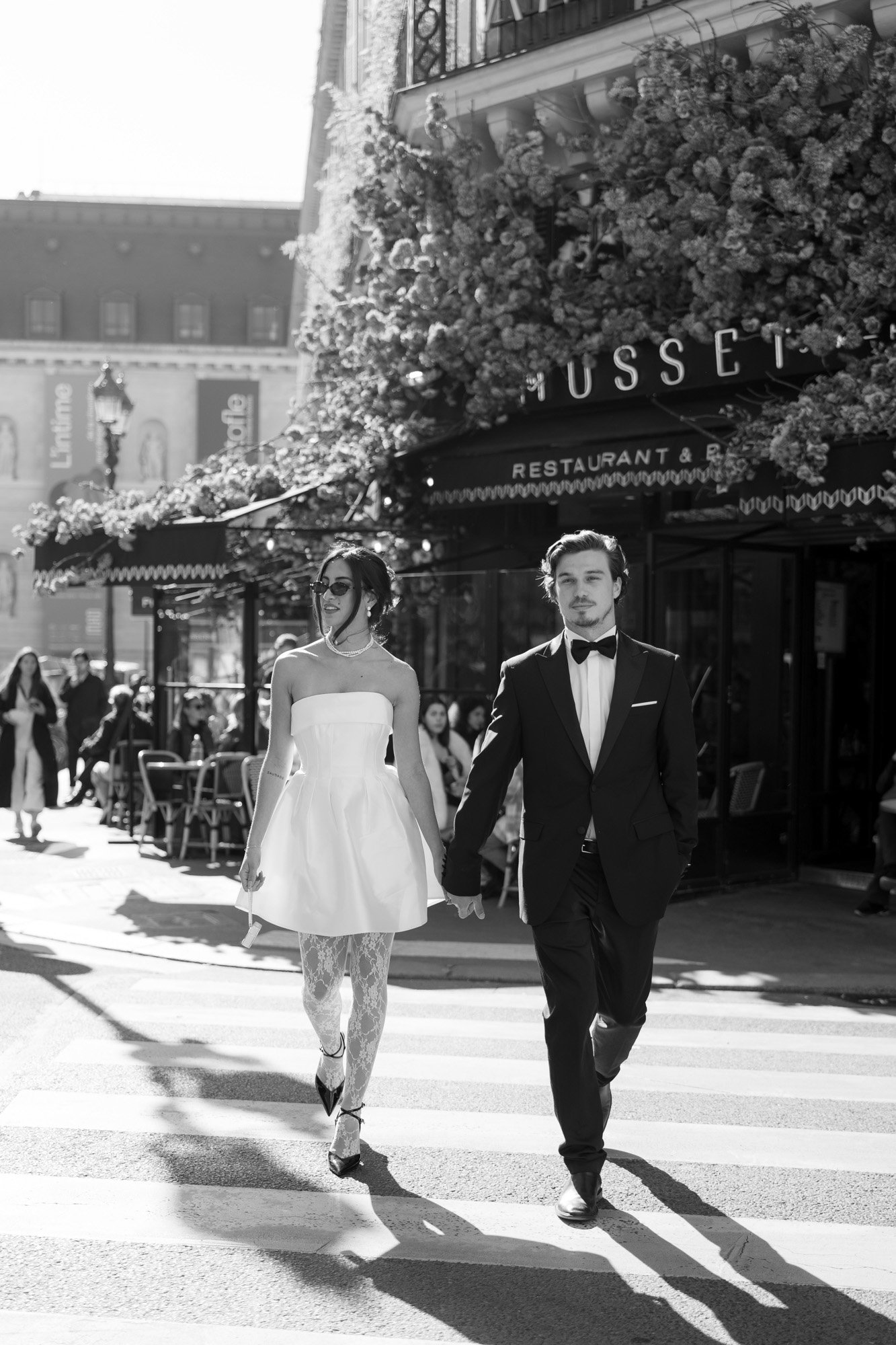 A couple dressed in formal attire holds hands while crossing a street in front of a restaurant, with people and outdoor seating in the background.