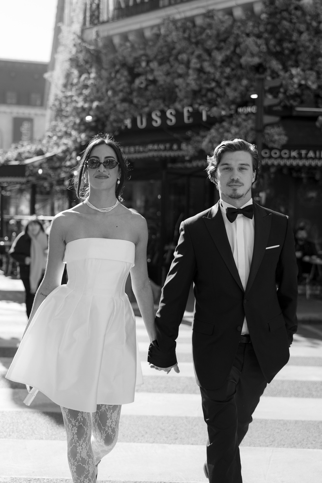 A woman in a strapless white dress and a man in a black tuxedo hold hands while walking on a city street in daylight.