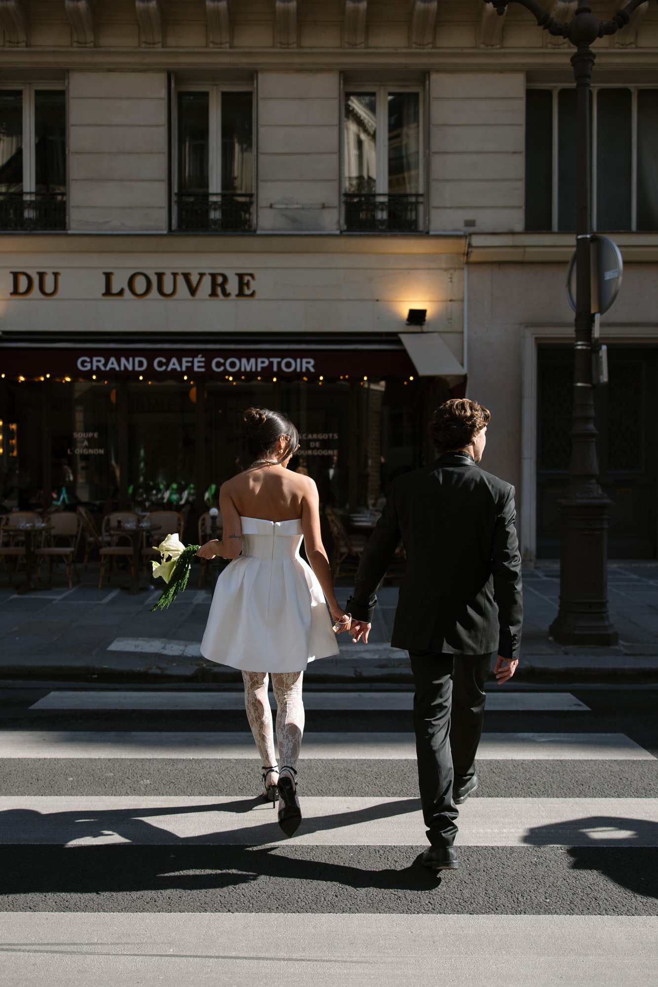 A woman in a white dress and a man in a suit hold hands while crossing a street in front of a café.