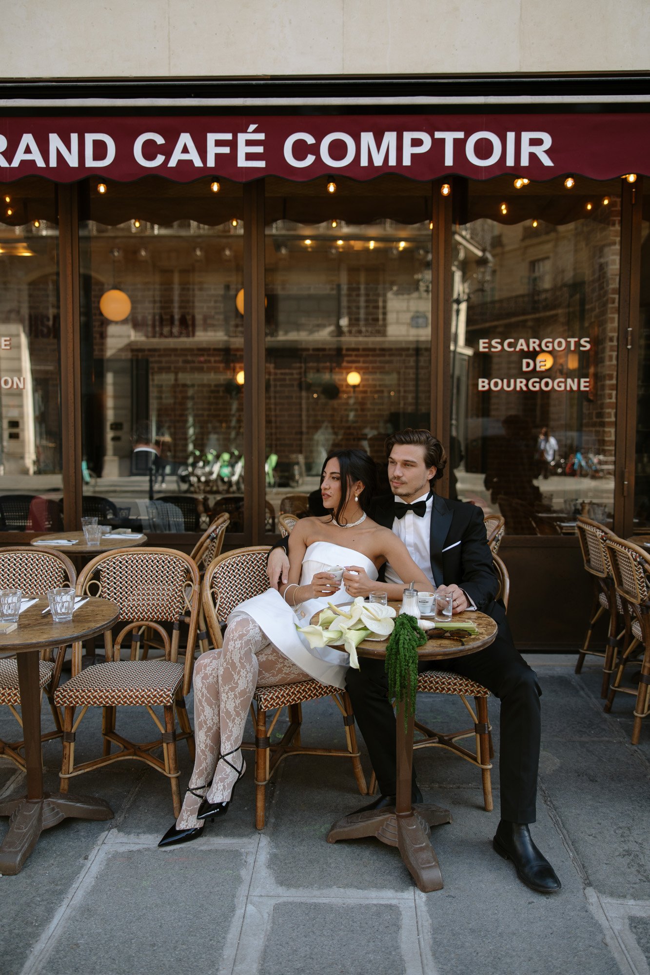 A woman in a white dress and a man in a black tuxedo sit together at an outdoor café table, with empty glasses and a plate of vegetables in front of them.