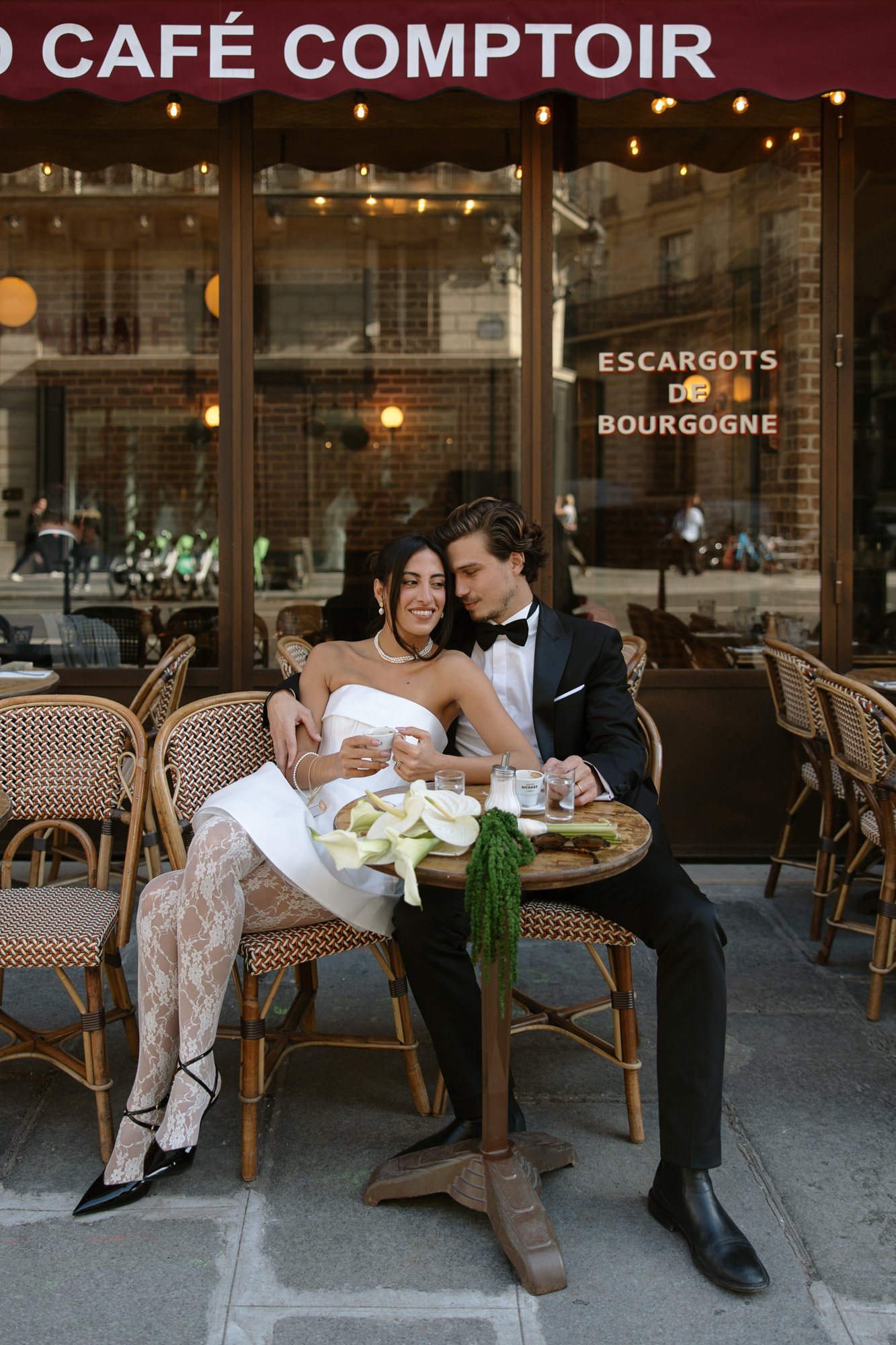 A couple dressed in formal attire sits closely together at an outdoor café table with food and drinks in front of them.