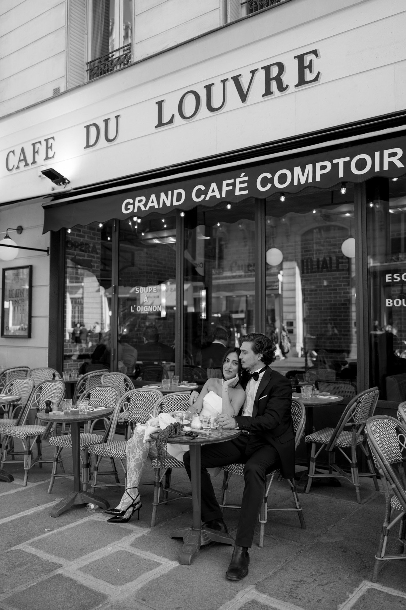 A couple dressed formally sits closely together at a small table outside Café du Louvre, with empty chairs around them on the terrace.