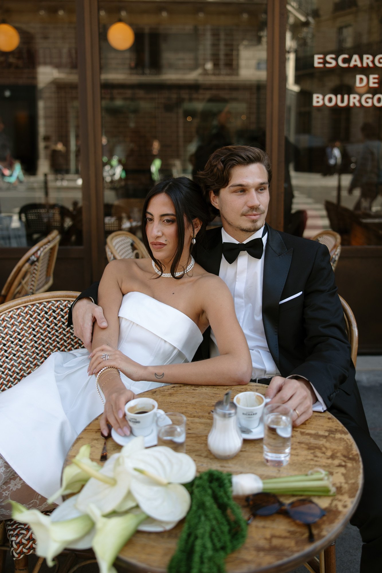 A couple dressed in formal attire sits at an outdoor café table with coffee, water, and flowers.