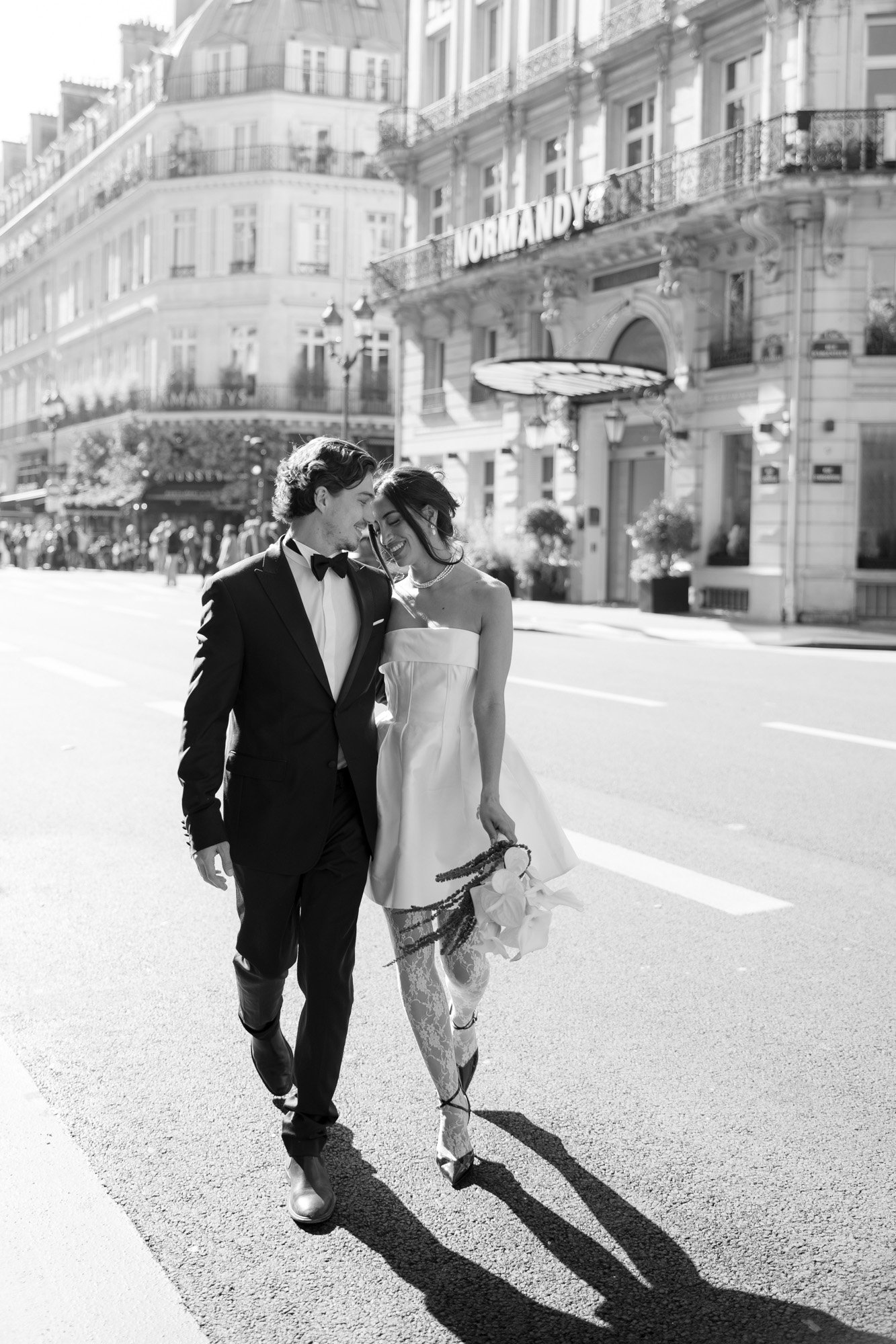 A couple dressed in formal attire walks closely together on a sunlit street in front of the Normandy Hotel.
