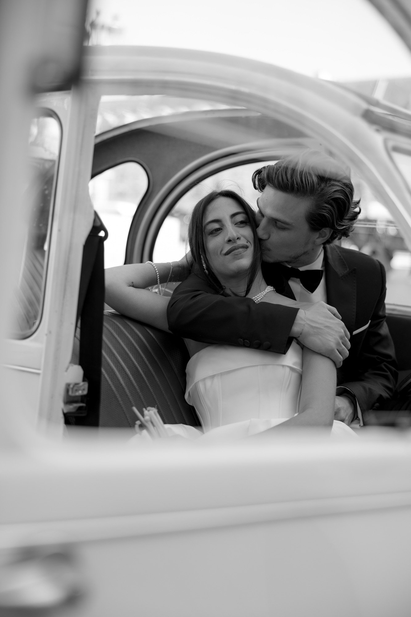 A couple in formal attire sits in the backseat of a car; the man kisses the womans cheek while she smiles and leans against him.