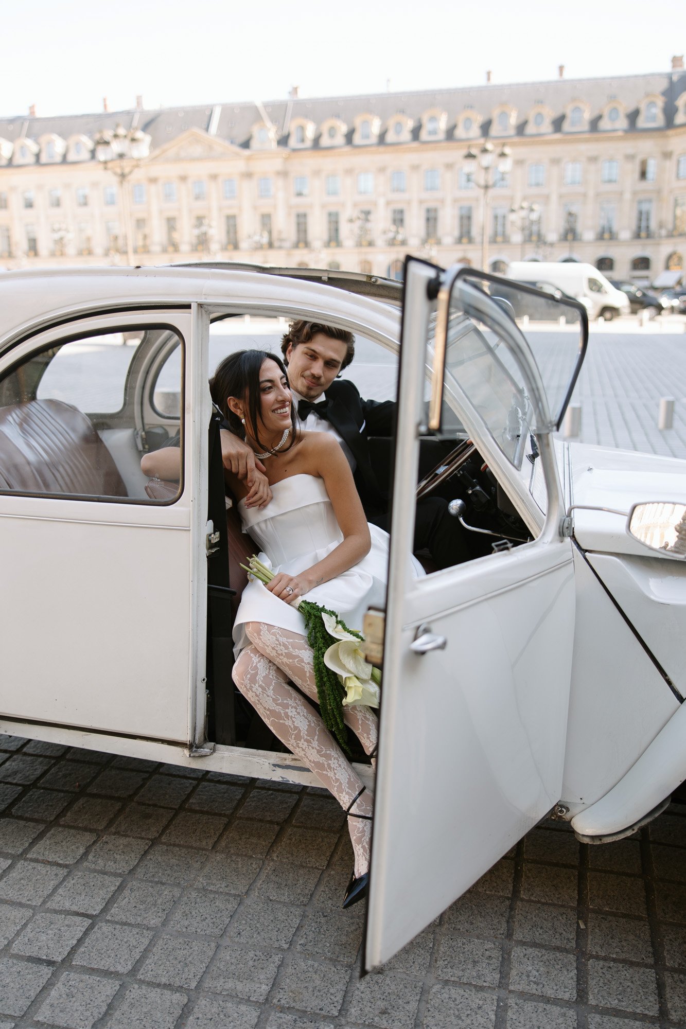 A bride and groom sit in a white vintage car with open doors, smiling at each other. Historic buildings and a paved plaza are visible in the background.