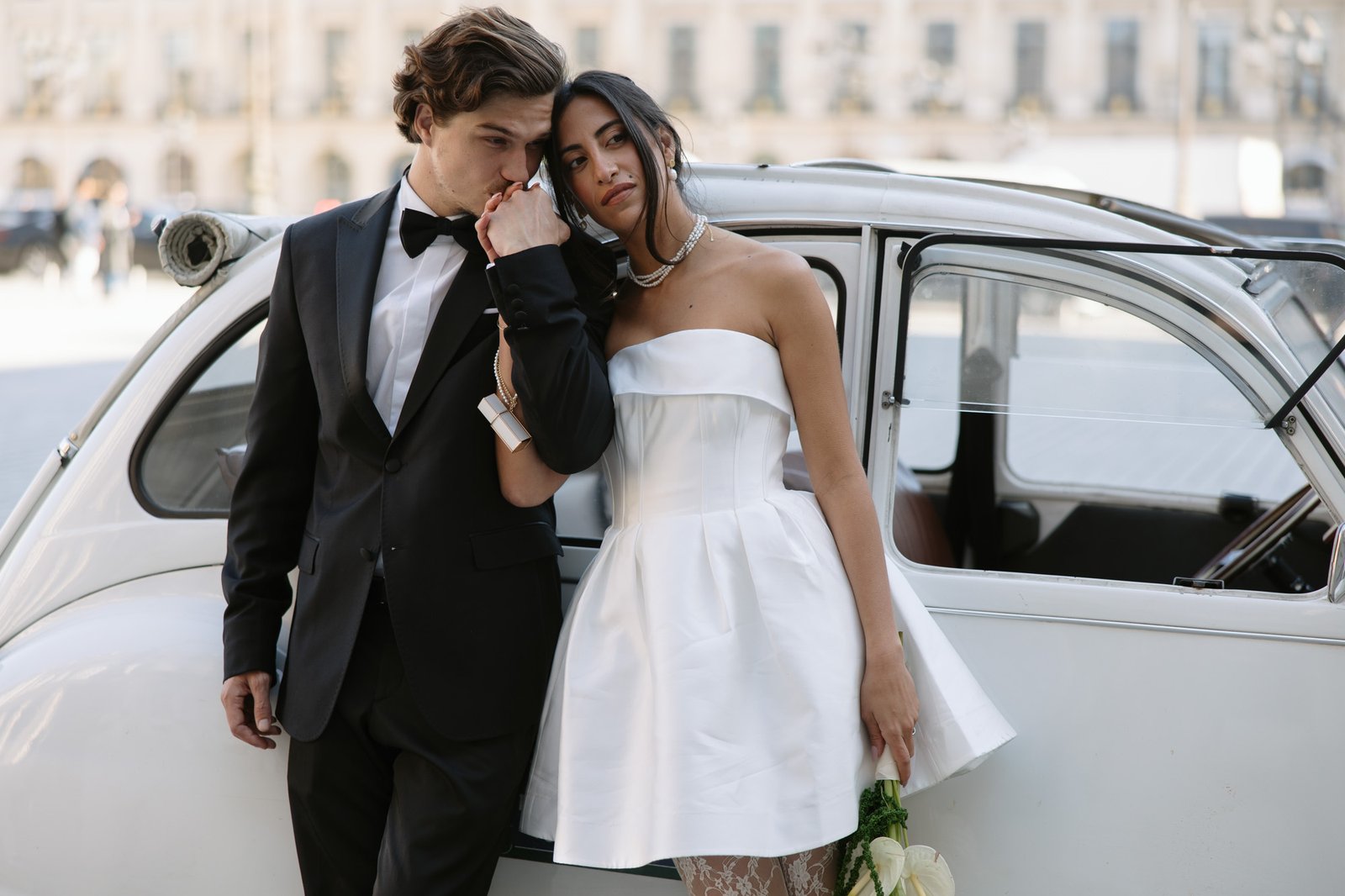 A man in a black suit kisses the hand of a woman in a white dress as they stand beside a vintage white car; she holds a bouquet of white flowers and leans against him.