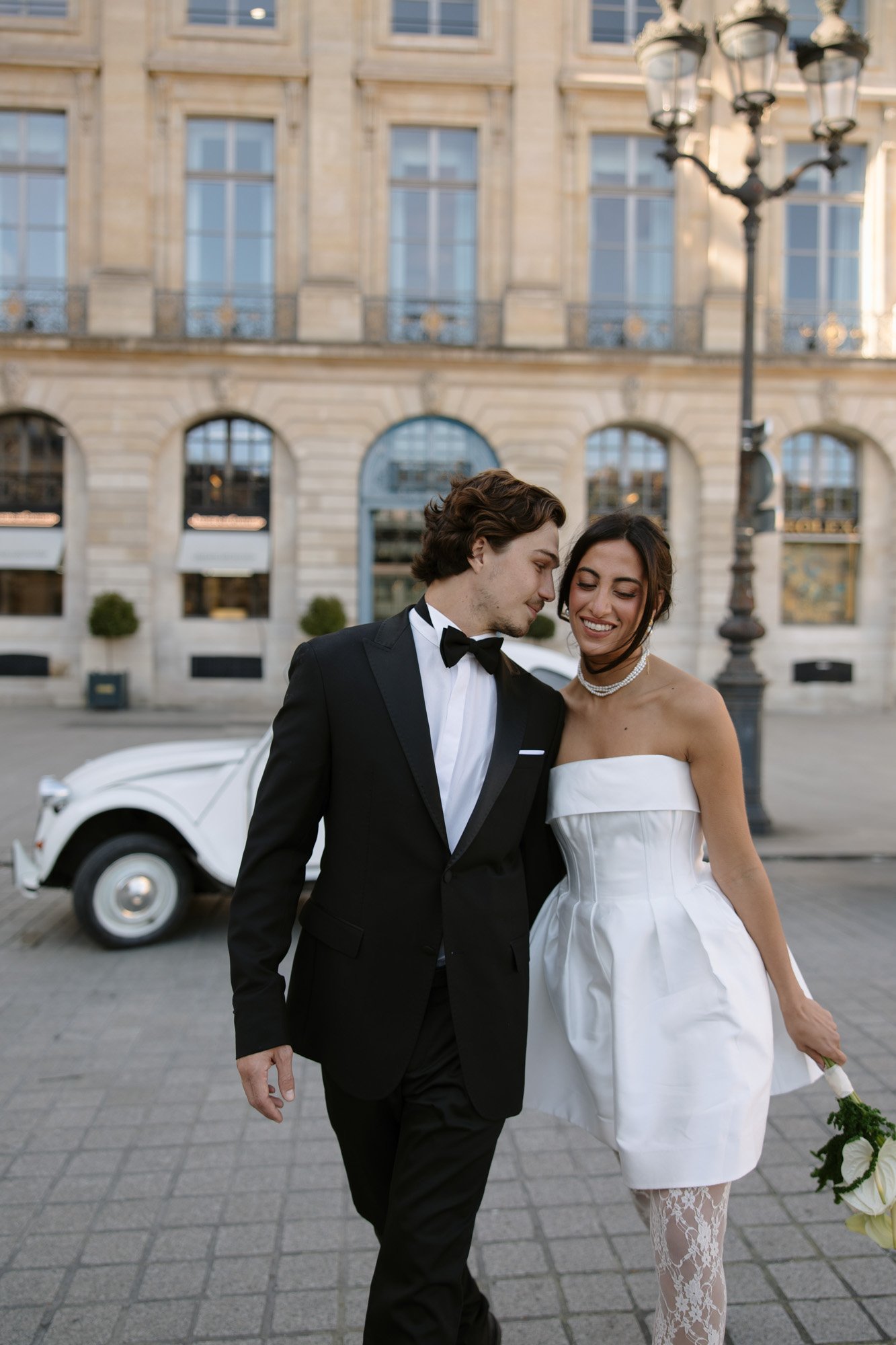 A couple dressed in formal wedding attire walks outside near a vintage white car and a historic building.