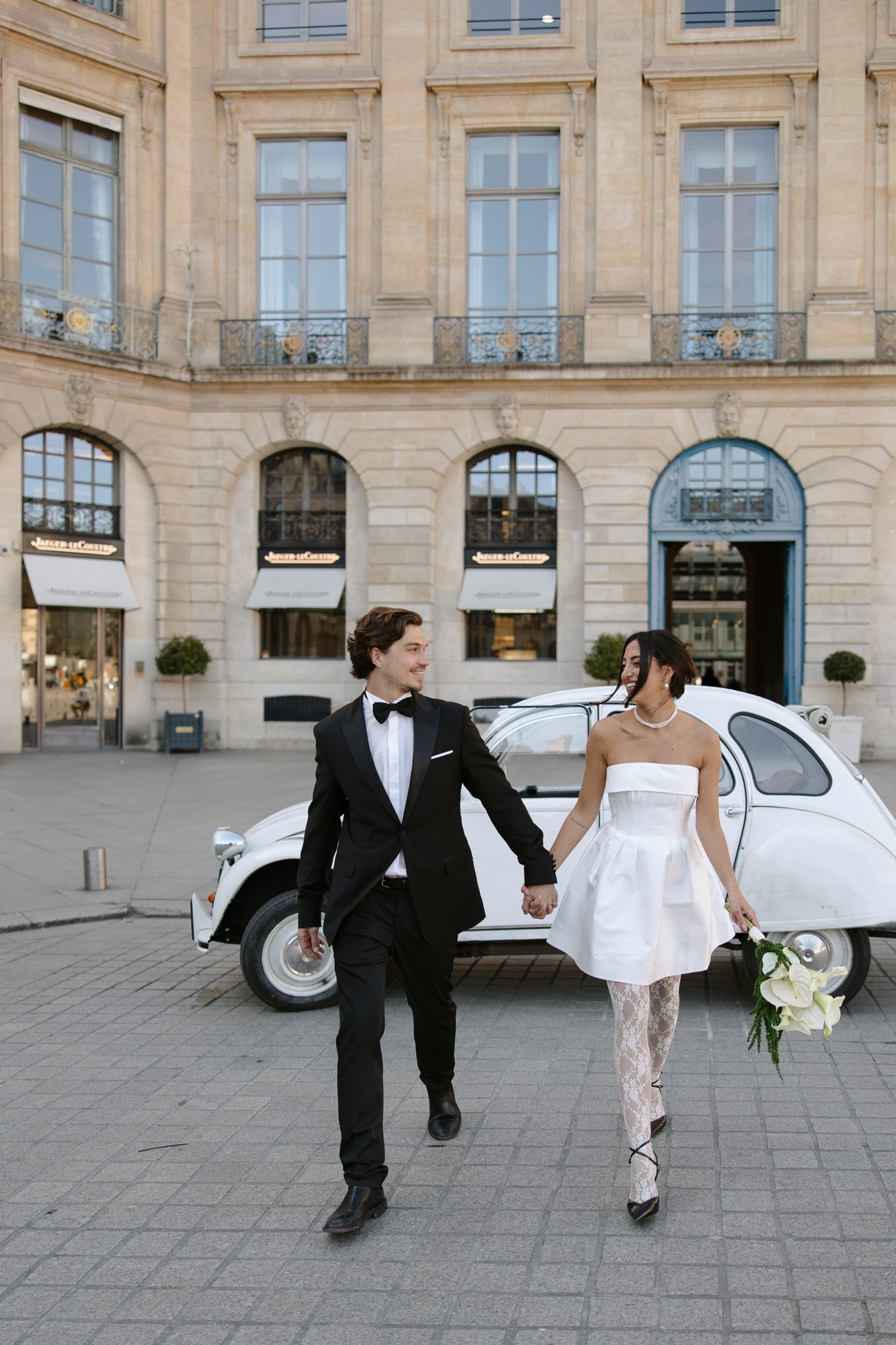 A couple dressed in formal attire, with the woman holding a bouquet, walk hand in hand in front of a classic white car outside a stone building. Paris wedding photographer.