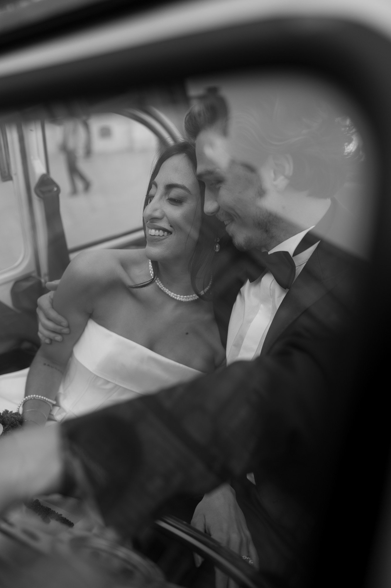 A couple dressed in formal attire sits closely together in a car, smiling and looking at each other through the window.