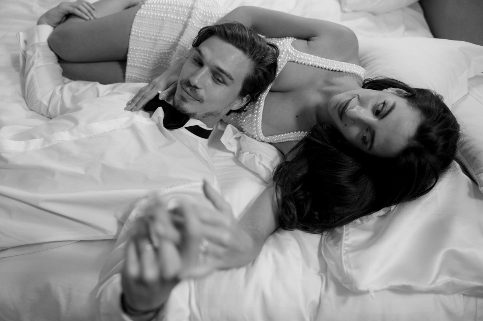 A man and a woman lie close together on a bed, holding hands and looking relaxed. The image is in black and white.