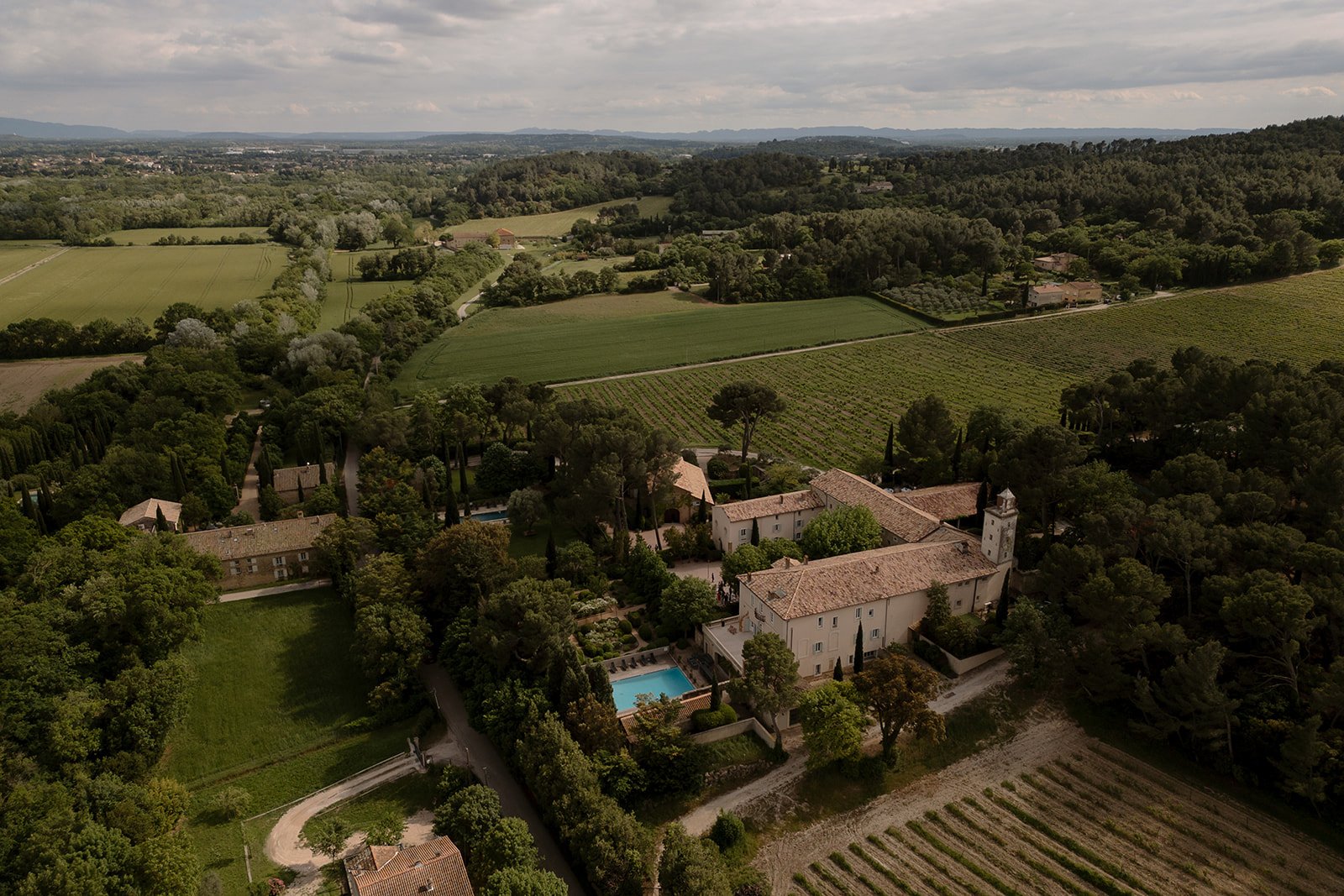 Aerial view of a large estate with tiled-roof buildings, a swimming pool, and surrounding vineyards and farmland under a cloudy sky. An elegant french wedding.