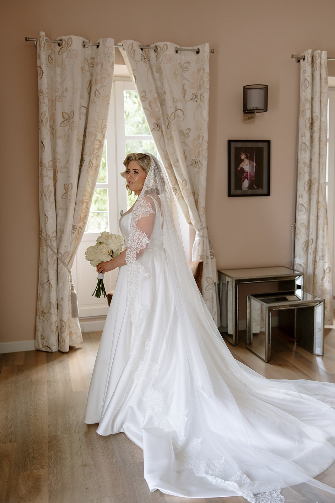 A bride in a long-sleeved white wedding dress and veil holds a bouquet of white flowers, standing indoors near large windows with light curtains. An elegant french wedding.
