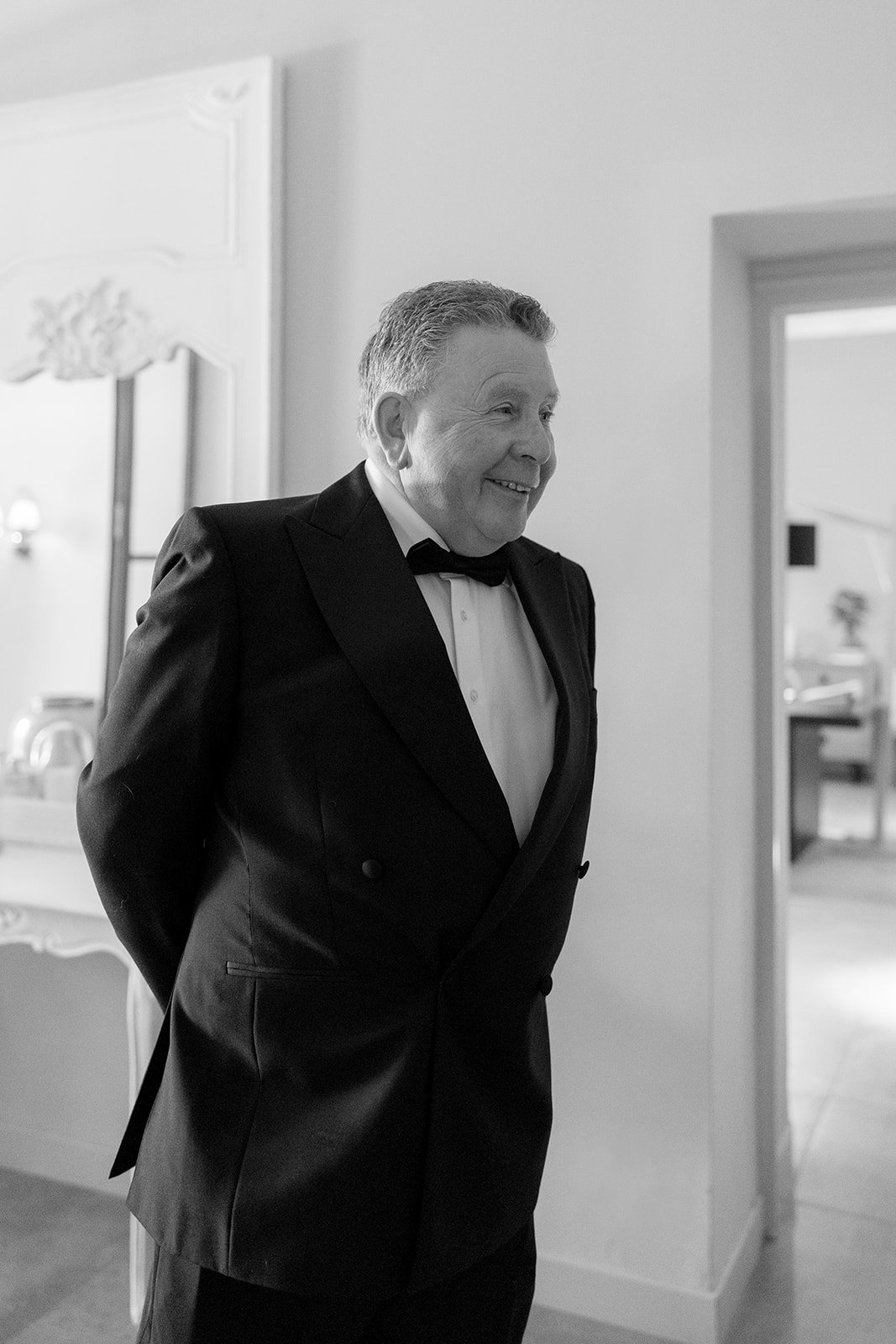 An older man in a tuxedo stands indoors, smiling with his hands behind his back. The setting appears to be a well-lit room with elegant decor.