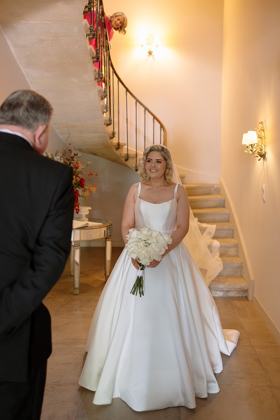 A bride in a white gown holding a bouquet stands at the bottom of a staircase, while a man in a suit faces her and a woman looks down from the stairs above.