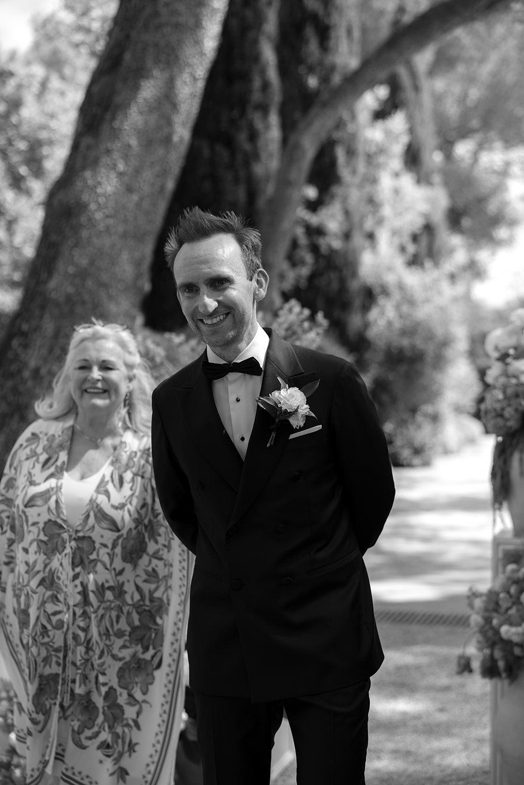 A man in a tuxedo, smiling, stands outdoors next to a woman in a patterned shawl. Trees and greenery are visible in the background. The image is in black and white.