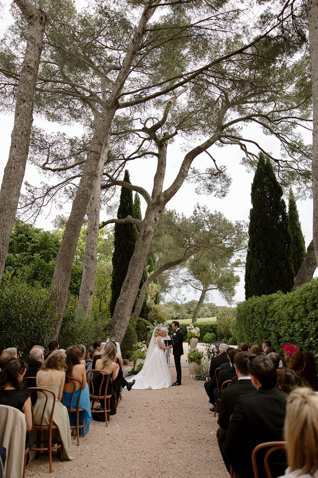 A bride and groom stand at the altar during an outdoor wedding ceremony surrounded by tall trees and seated guests. An elegant french wedding.