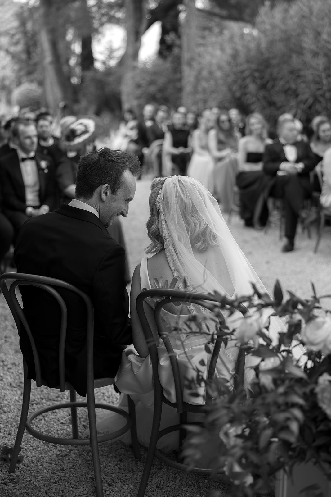 A bride and groom sit close together at an outdoor wedding ceremony, facing guests seated in rows, surrounded by greenery. An elegant french wedding.