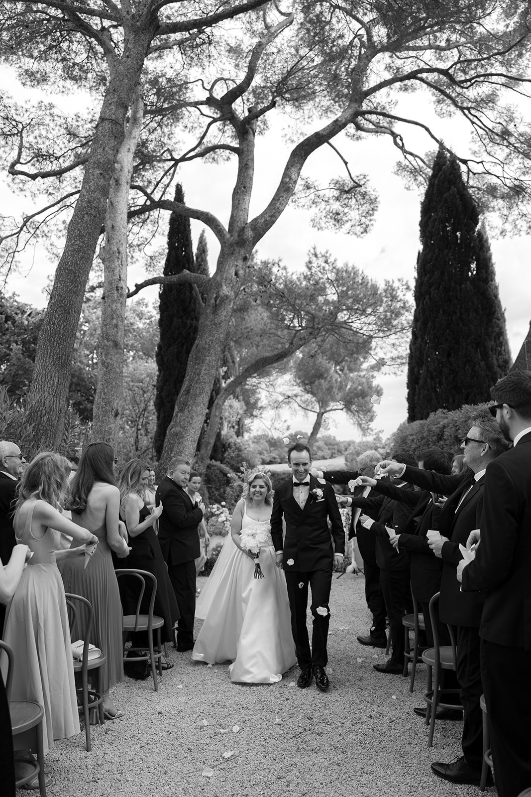 A bride and groom walk down an outdoor aisle as guests seated on either side applaud and take photos, surrounded by tall trees. An elegant french wedding.