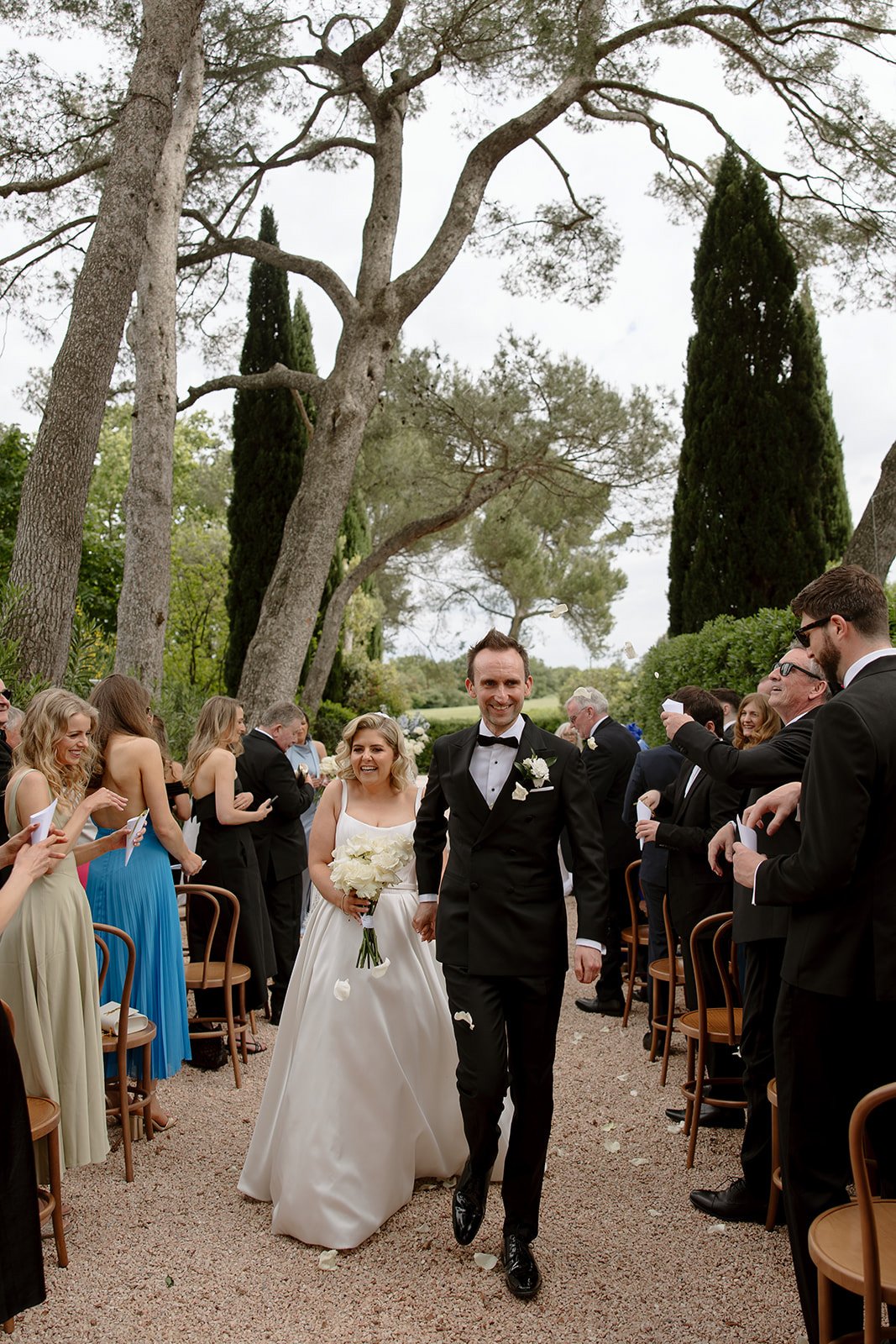 A newlywed couple walks down an outdoor aisle lined with wooden chairs, surrounded by guests in formal attire and tall trees. An elegant french wedding.