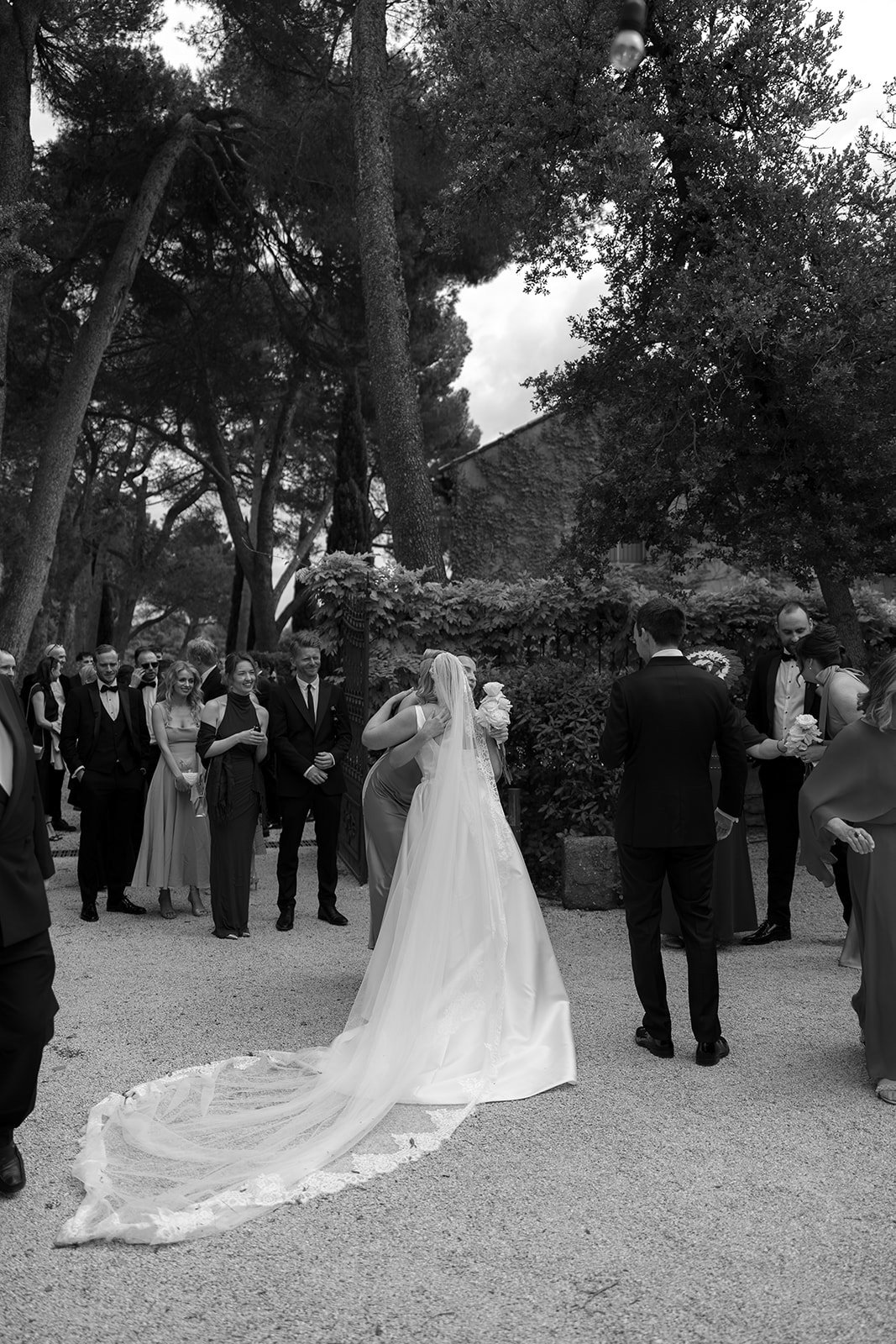 A bride in a long gown and veil hugs a woman outdoors, surrounded by guests in formal attire, while a man stands nearby. An elegant french wedding.