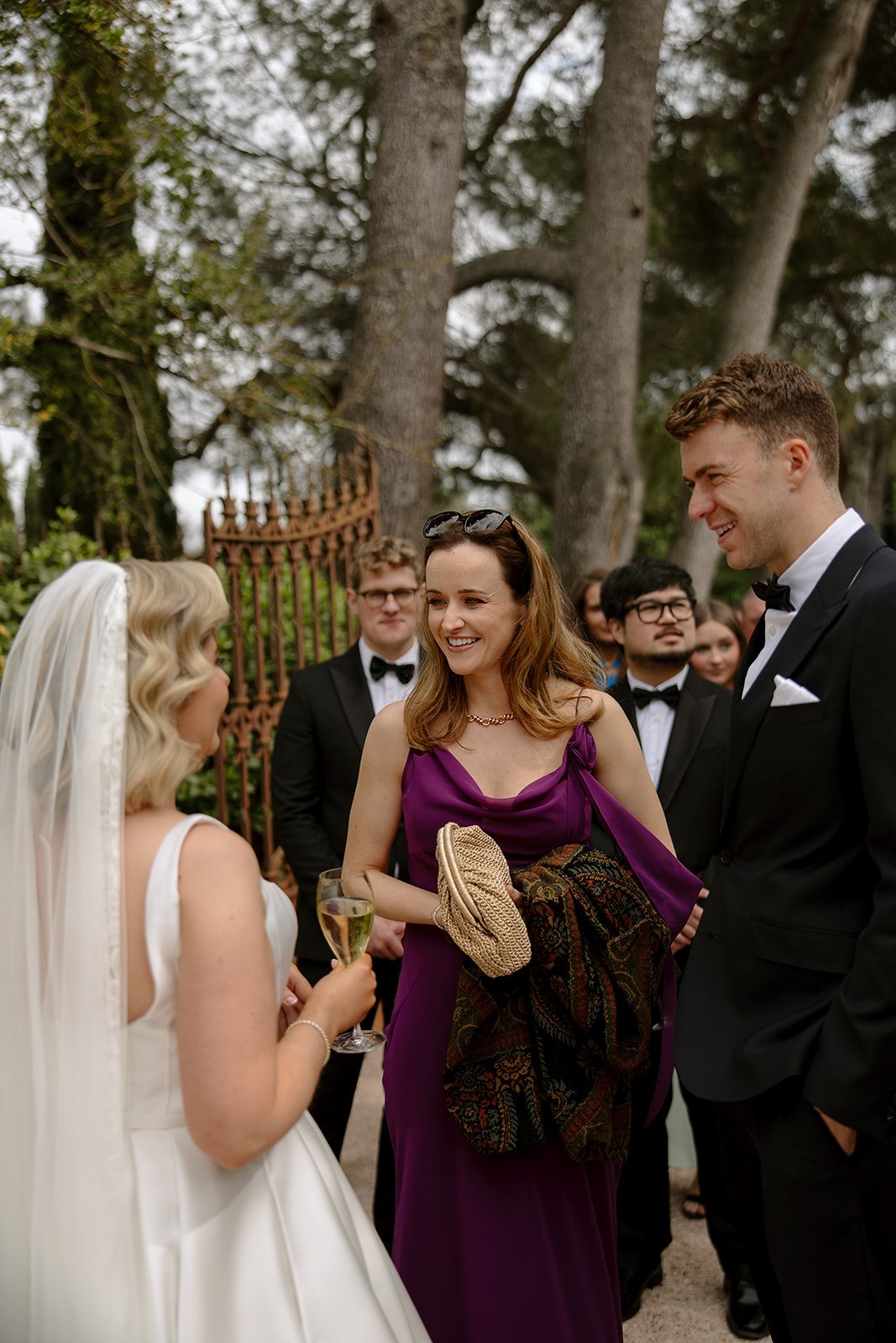 A bride in a white dress with a veil talks to guests in formal attire at an outdoor event, with trees and an iron fence in the background. An elegant french wedding.