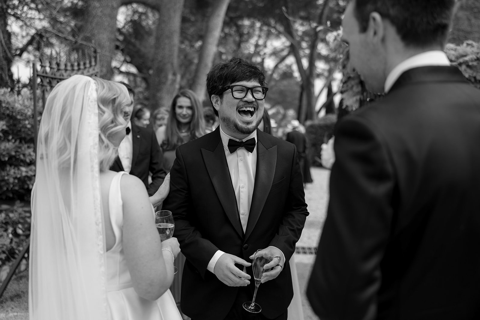 A man in a tuxedo laughs while holding a drink, standing outside with a bride and another guest at what appears to be a wedding reception.
