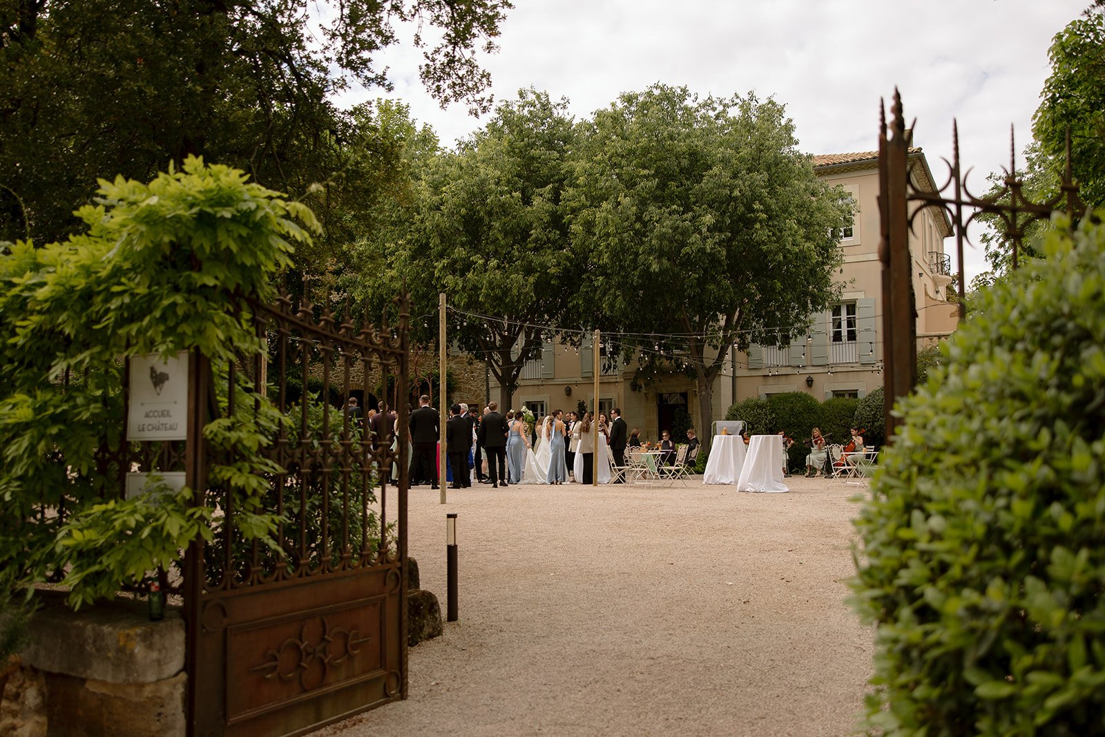 A group of people gather outdoors near a large house, surrounded by trees and greenery, with tables and string lights set up for an event.