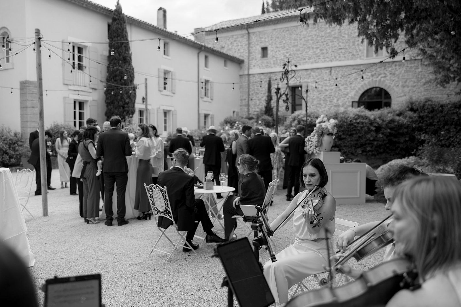 A group of people socializes outdoors at a formal event, while a small string ensemble performs in the foreground.