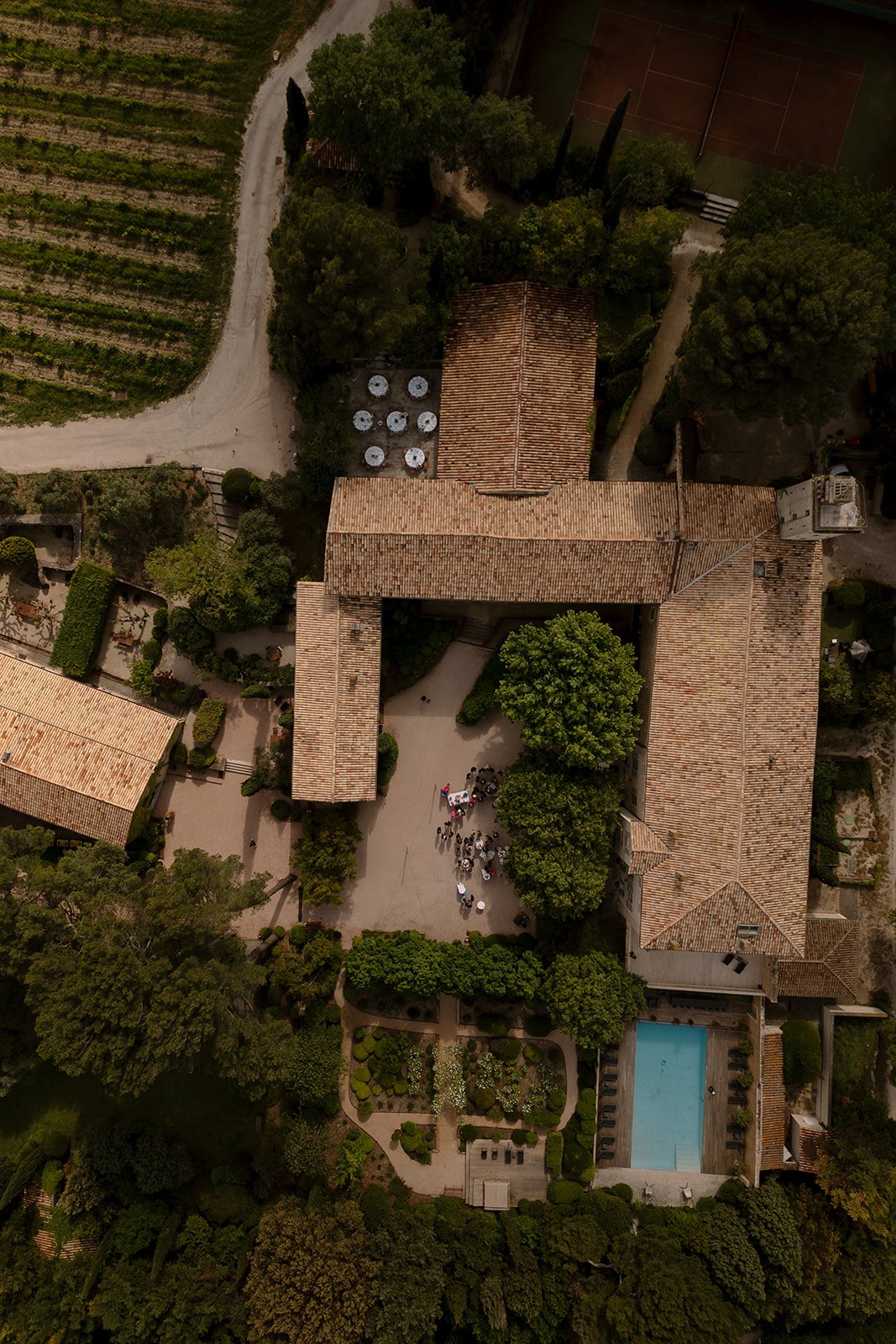 Aerial view of a rustic estate with tiled roofs, a courtyard with people seated at tables, a swimming pool, tennis court, gardens, and adjacent vineyard.