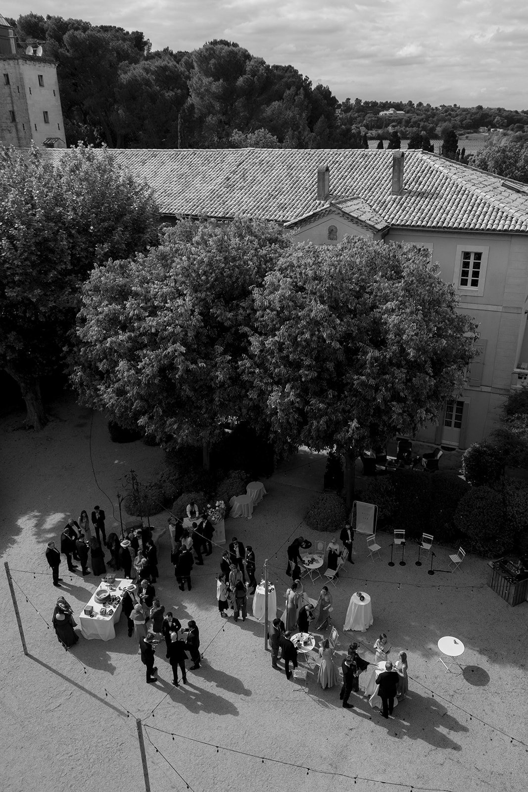 A group of people gather around tables set up outdoors in a courtyard, shaded by trees, with a large building and trees in the background.