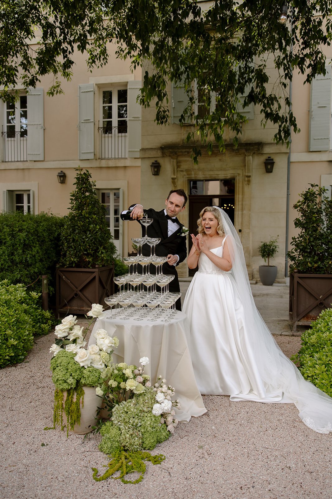 A bride and groom stand outside a building as the groom pours champagne into a pyramid of glasses, with the bride looking surprised. Floral arrangements decorate the scene. An elegant french wedding.