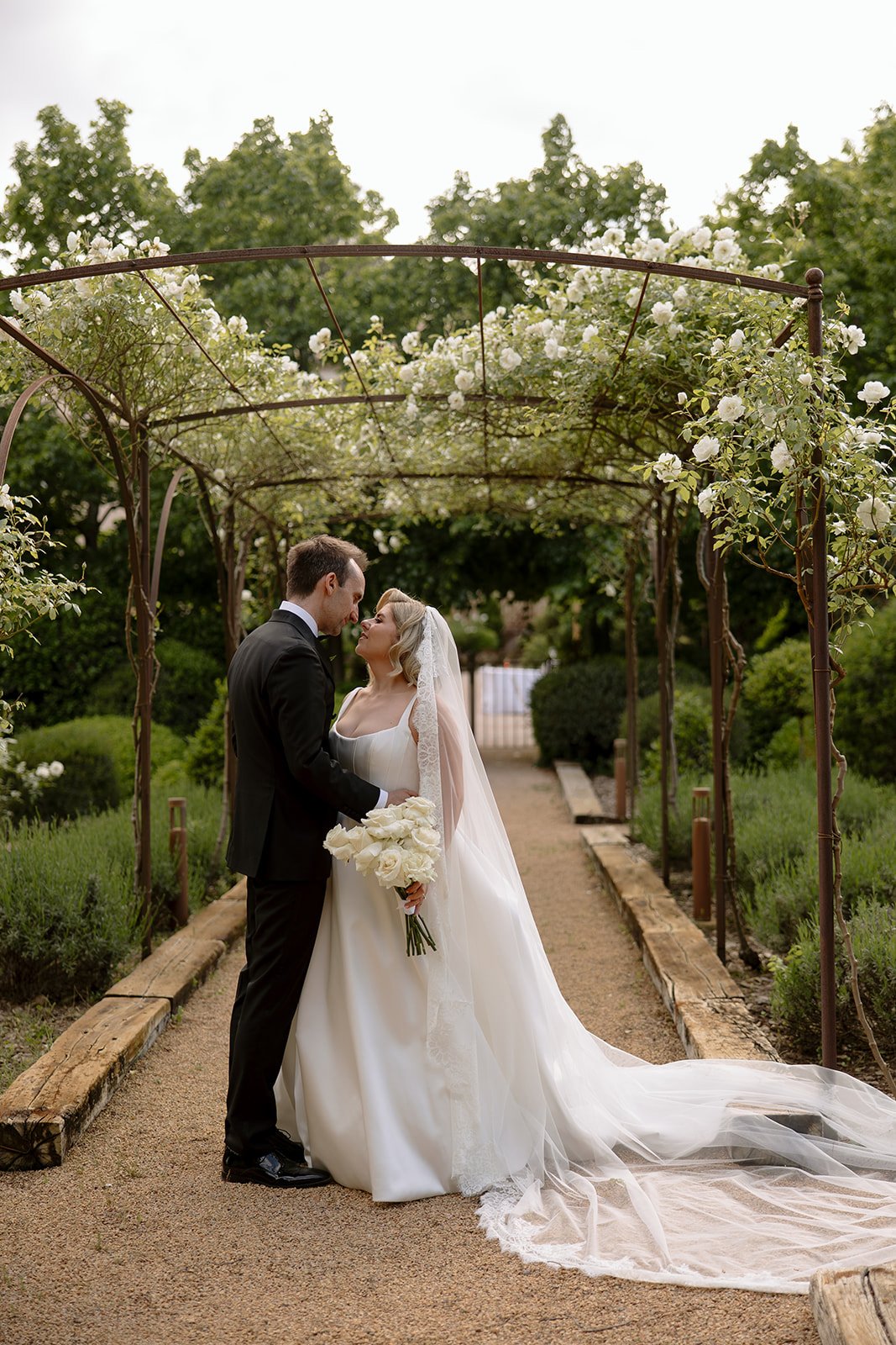 A bride and groom stand under a floral archway in a garden, holding a bouquet of white flowers and sharing an intimate moment on their wedding day. An elegant french wedding.