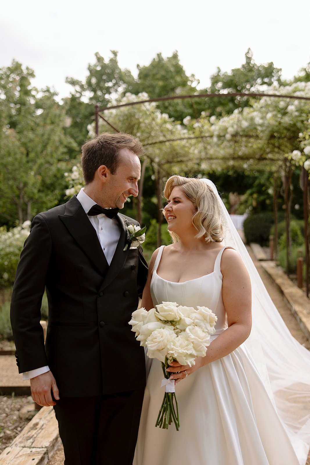A bride in a white dress holding a bouquet of white roses stands beside a groom in a black tuxedo; they are smiling at each other outdoors.