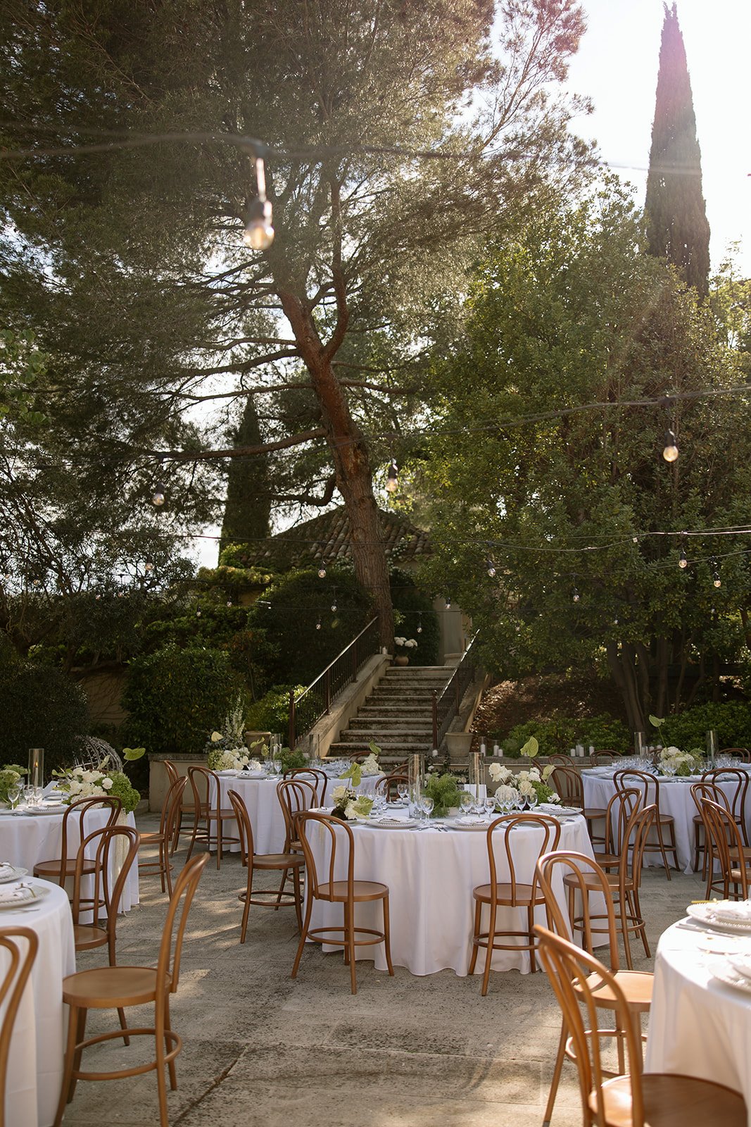 Outdoor dining area with round tables covered in white tablecloths, wooden chairs, and string lights, set amid trees and greenery near stone steps. An elegant french wedding.