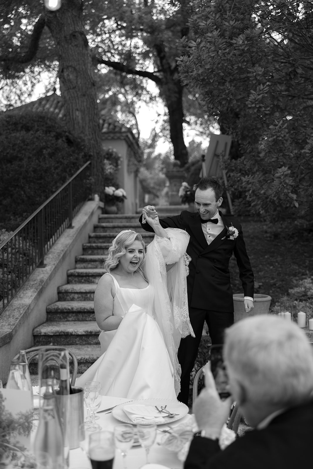 A bride sits and smiles while a groom holds her hand and lifts her arm; they are outdoors near a staircase, with another person in the foreground.