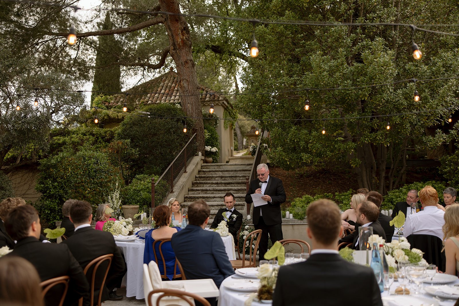 An outdoor wedding reception with guests seated at tables listening to a man giving a speech near stairs, surrounded by greenery and string lights.