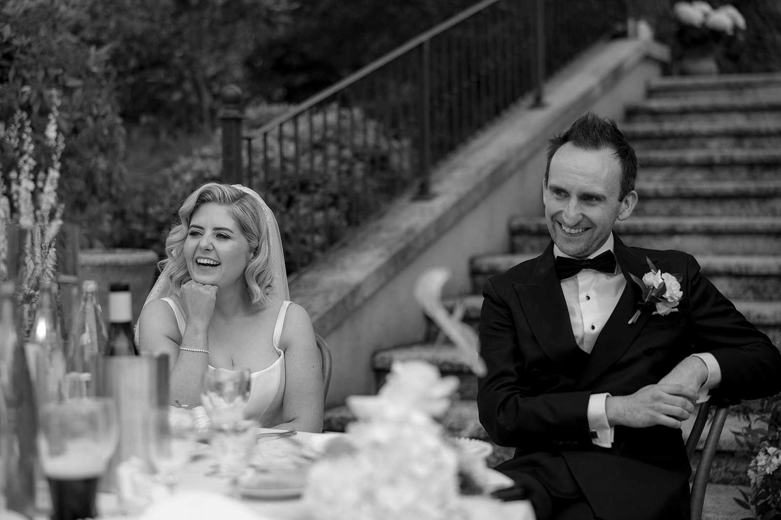 A bride and groom sit at an outdoor table, smiling and dressed in wedding attire, with stone steps and greenery in the background.