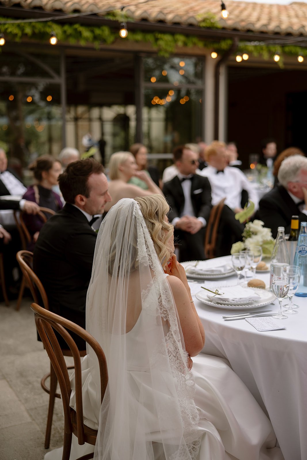A bride in a white dress and veil sits at a table during an outdoor wedding reception, surrounded by formally dressed guests.