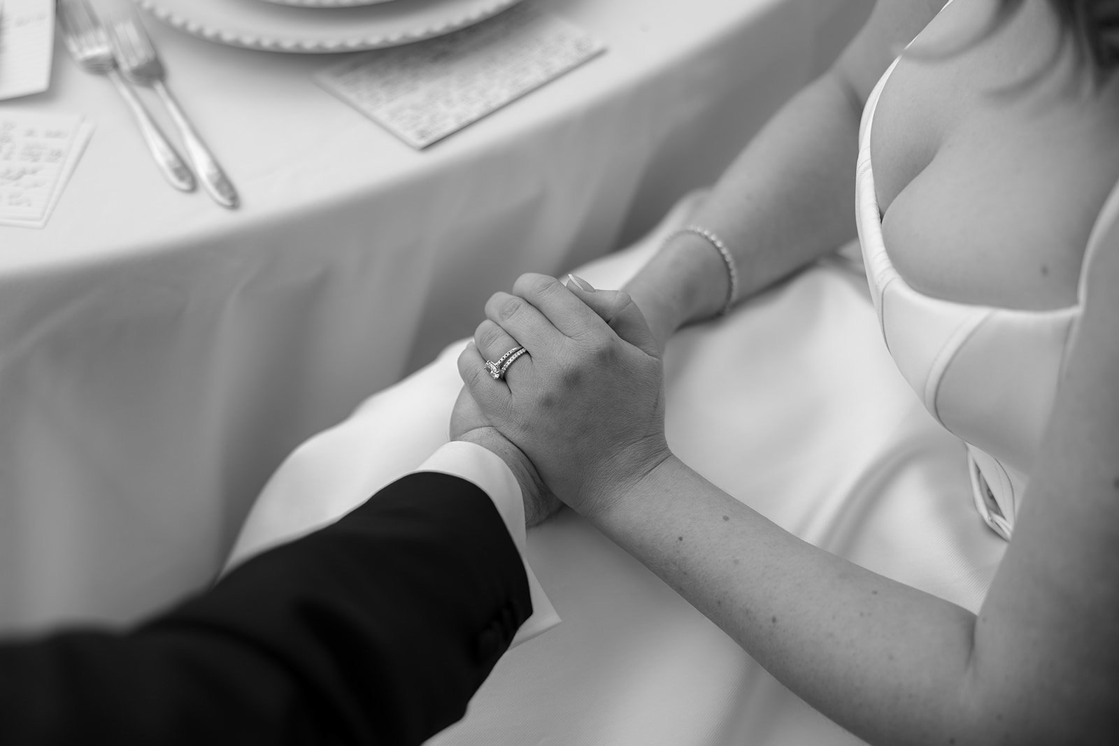 A close-up of a couple holding hands at a table, with the woman wearing a wedding dress and a ring set on her finger.