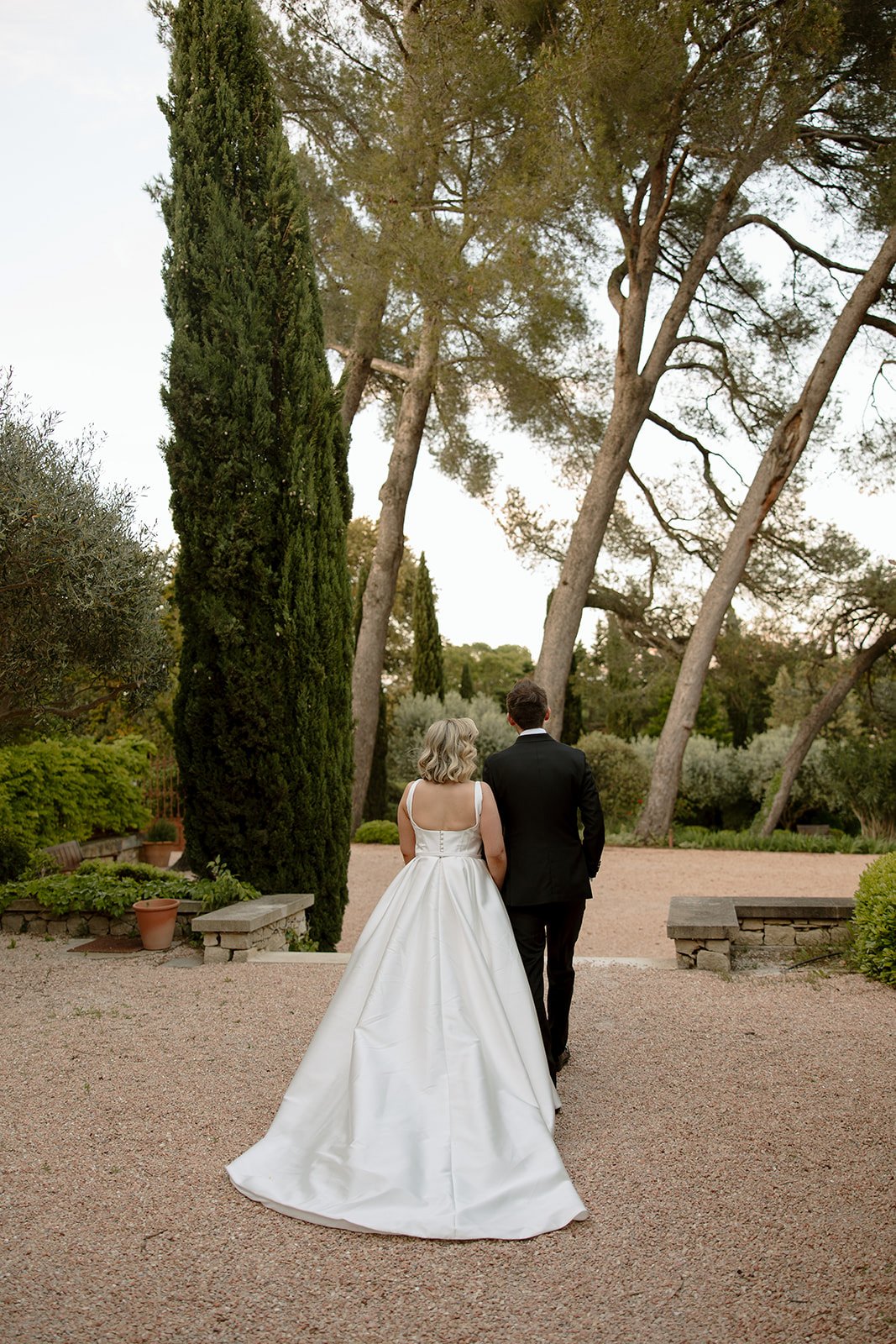 A bride in a white gown and a groom in a black suit walk together on a gravel path in a garden surrounded by tall trees and greenery.