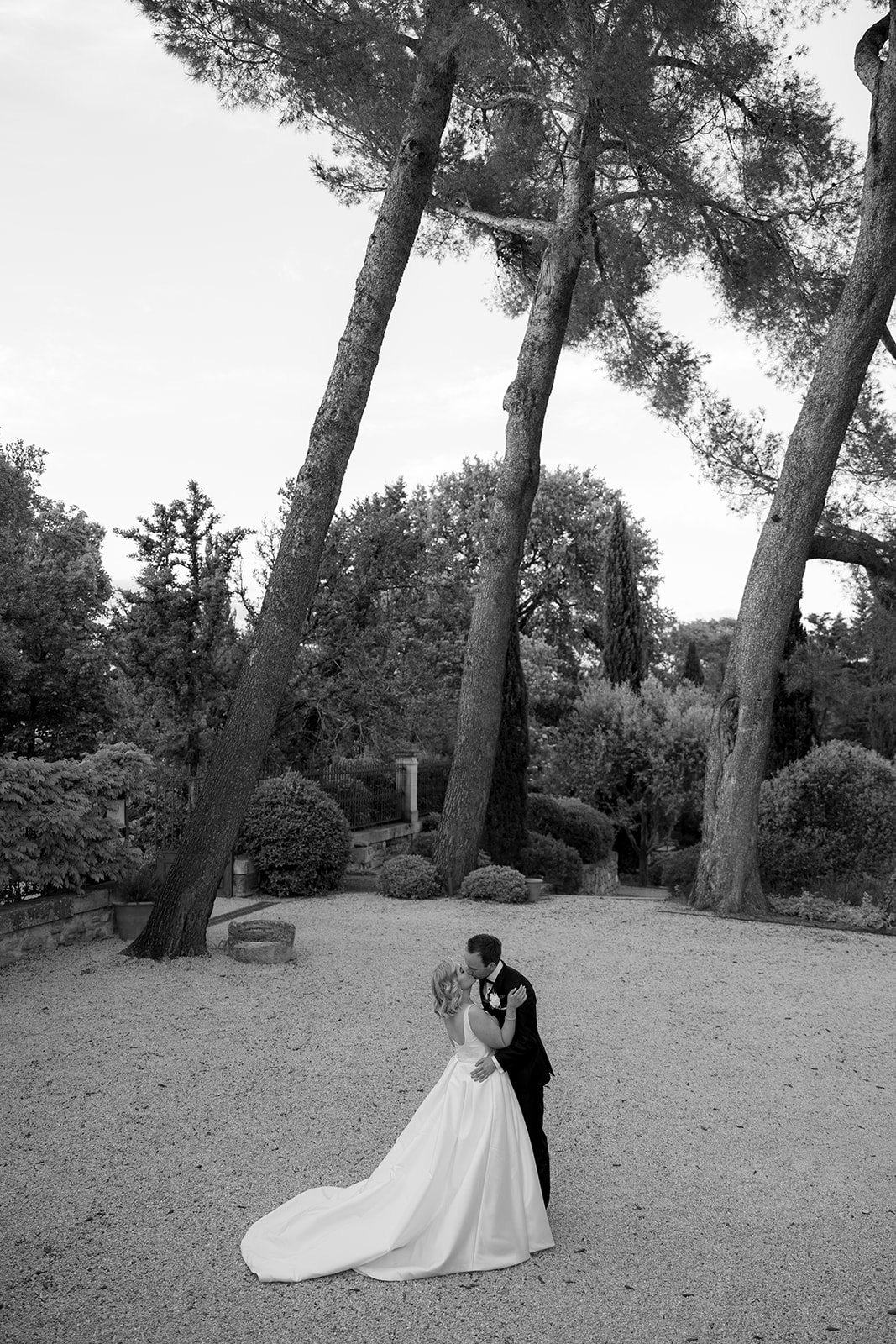 A bride and groom stand close together outdoors under tall trees, surrounded by greenery, on a gravel surface.