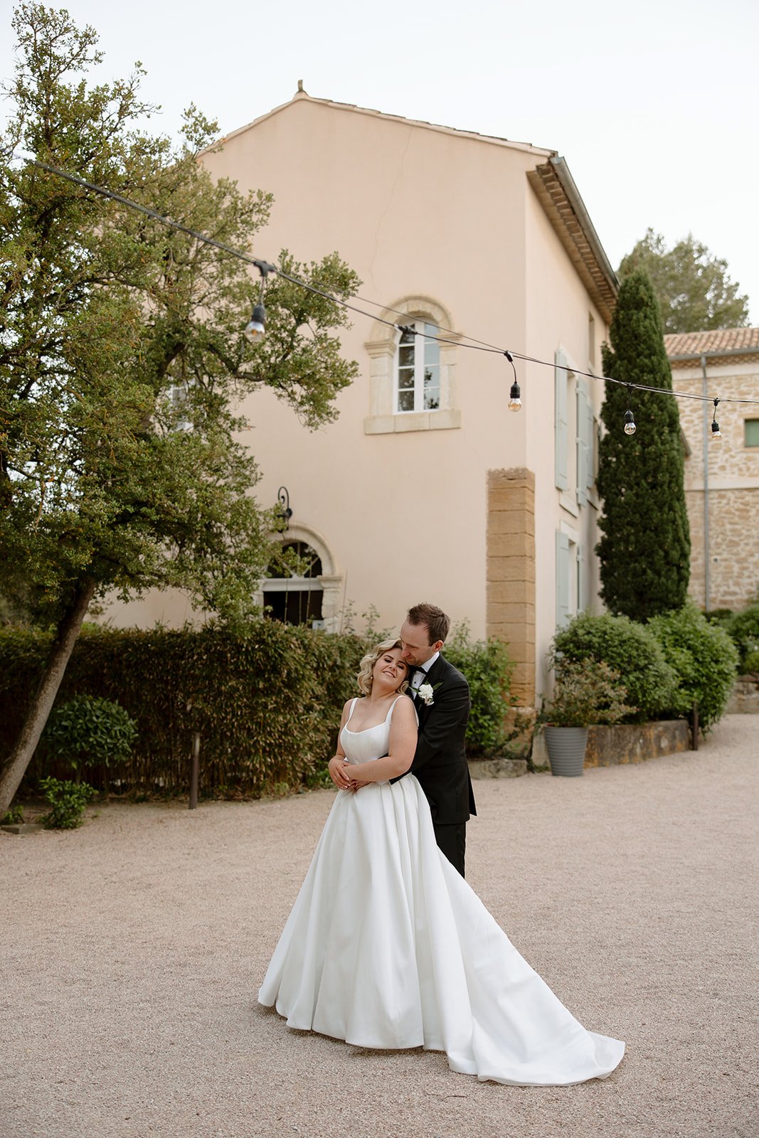 A bride in a white gown and a groom in a suit embrace outside a light-colored building with greenery and string lights in the background. An elegant french wedding.