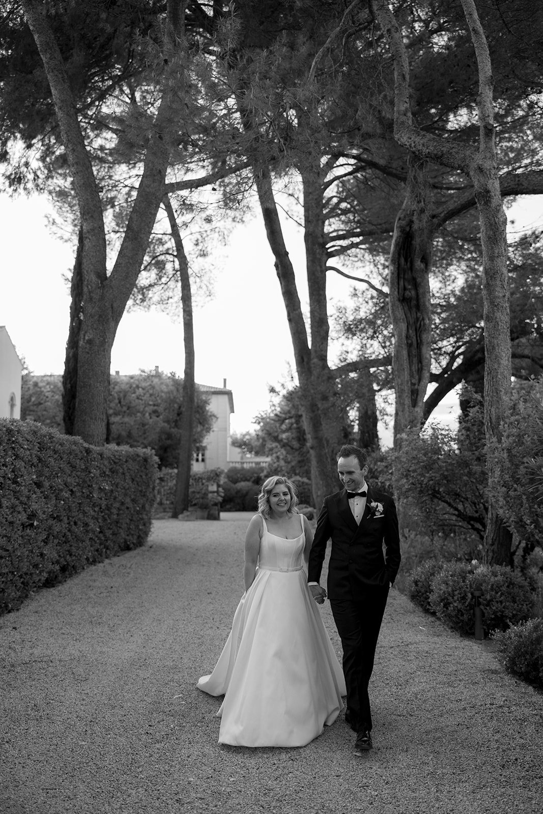A bride and groom walk hand in hand down a tree-lined path, dressed in formal wedding attire.