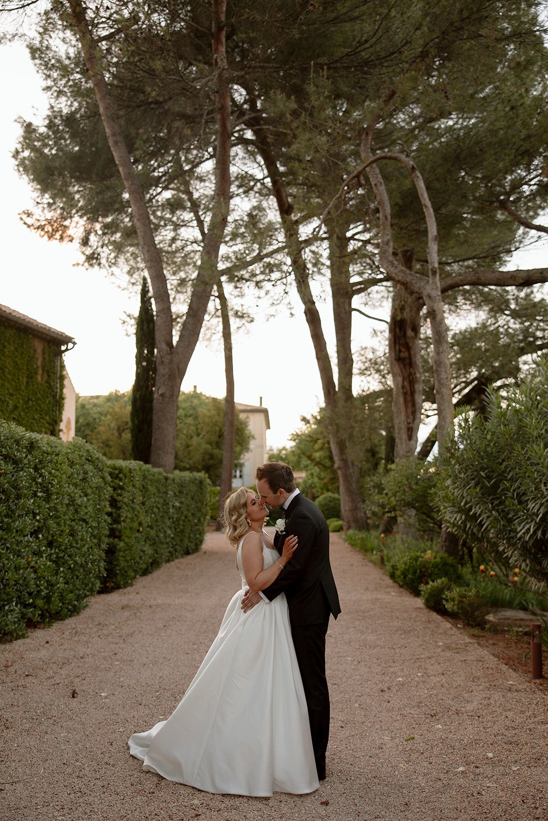 A bride and groom kiss on a tree-lined pathway, with greenery and large trees surrounding them. An elegant french wedding.